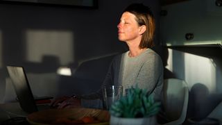 Woman practising meditation at home with laptop on table in a ray of sunlight