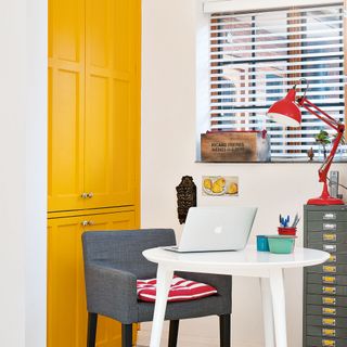 laptop on white table in kitchen with yellow cupboard door and red table lamp