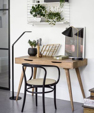 Contemporary desk area with wooden desk, black desk chair, sleek black floor lamp beside desk, table lamp, artwork and decorative ornaments on desk, white string shelf mounted above desk, decorated with plants, books and storage boxes