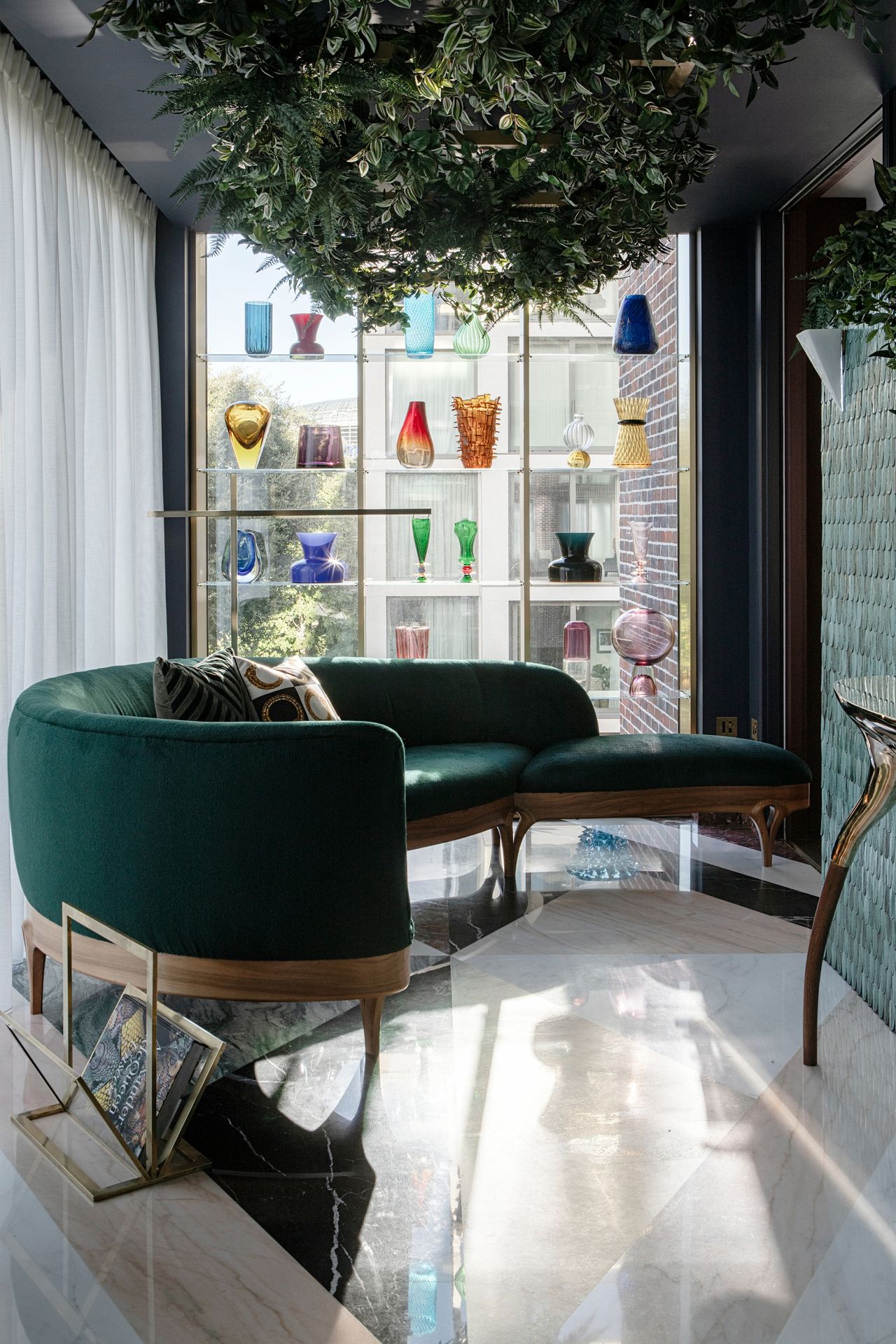 Image of an emerald green sitting room with lots of windows. Two of the walls are all window &mdash; one with a clear shelving unit with colorful vases on it, and the other with a white curtain covering the windows. There is a curved, dark green sofa with a triangle ottoman attached. There are plants hanging from the ceiling and the floors are a mixed marble &mdash; green, white, and pale pink marble in a geometric pattern.