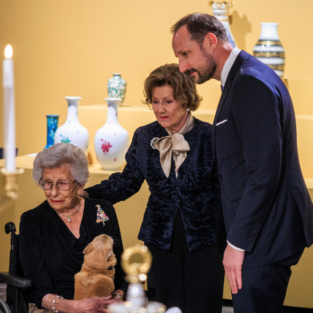 King Haakon, Queen Sonja and Princess Astrid, sitting in a wheelchair with her dog, viewing an exhibition 