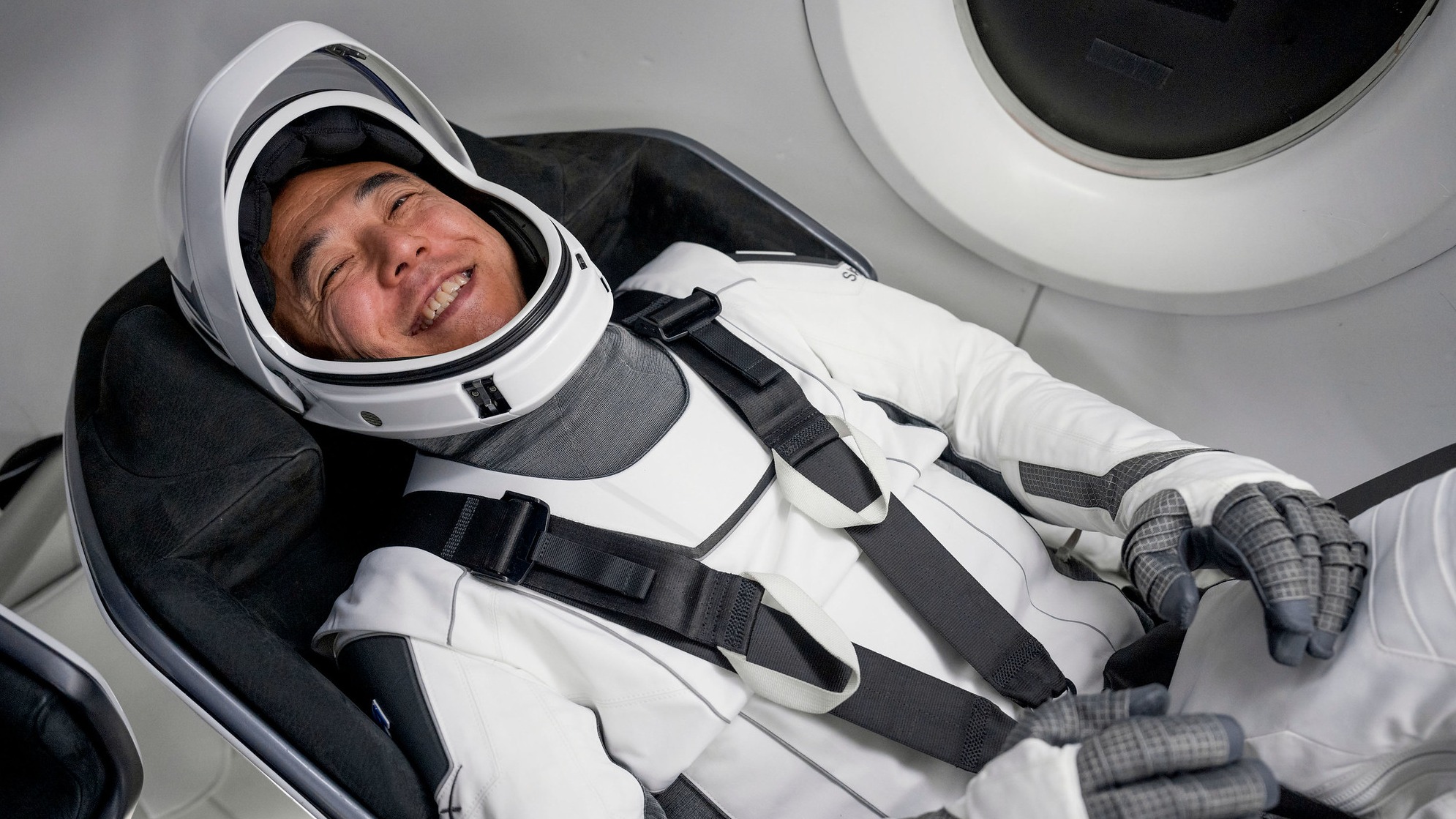 Satoshi Furukawa está acostado en un traje espacial en el asiento de la nave espacial y sonríe.  Aparece una ventana parcialmente circular en la parte superior derecha