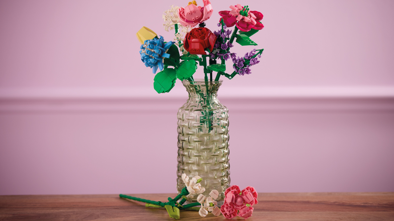 A bouquet of Aldi building block flowers on a wooden table in front of a pink background. There are roses, clover and lavendar in a clear ribbed vase.