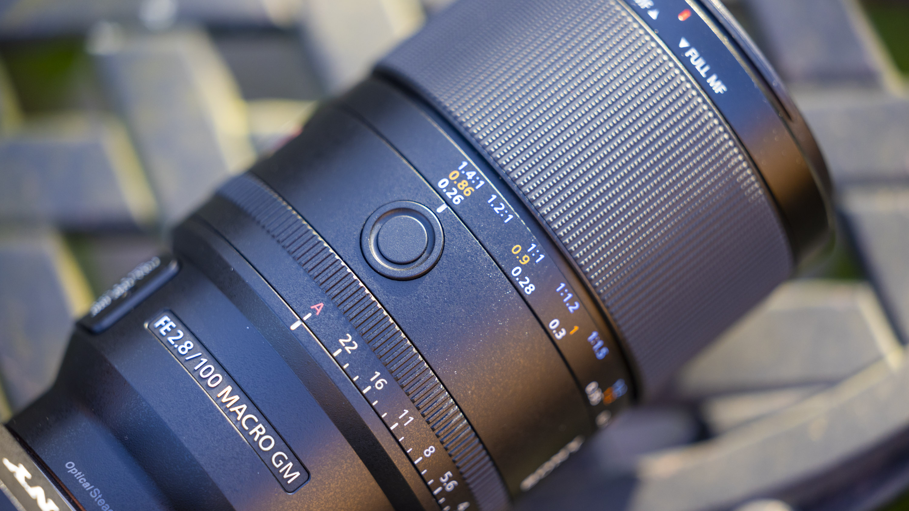 A product shot of the Sony FE 100mm f/2.8 Macro GM OSS attached to a Sony A7R IV outdoors on a metal garden table with a blurry background