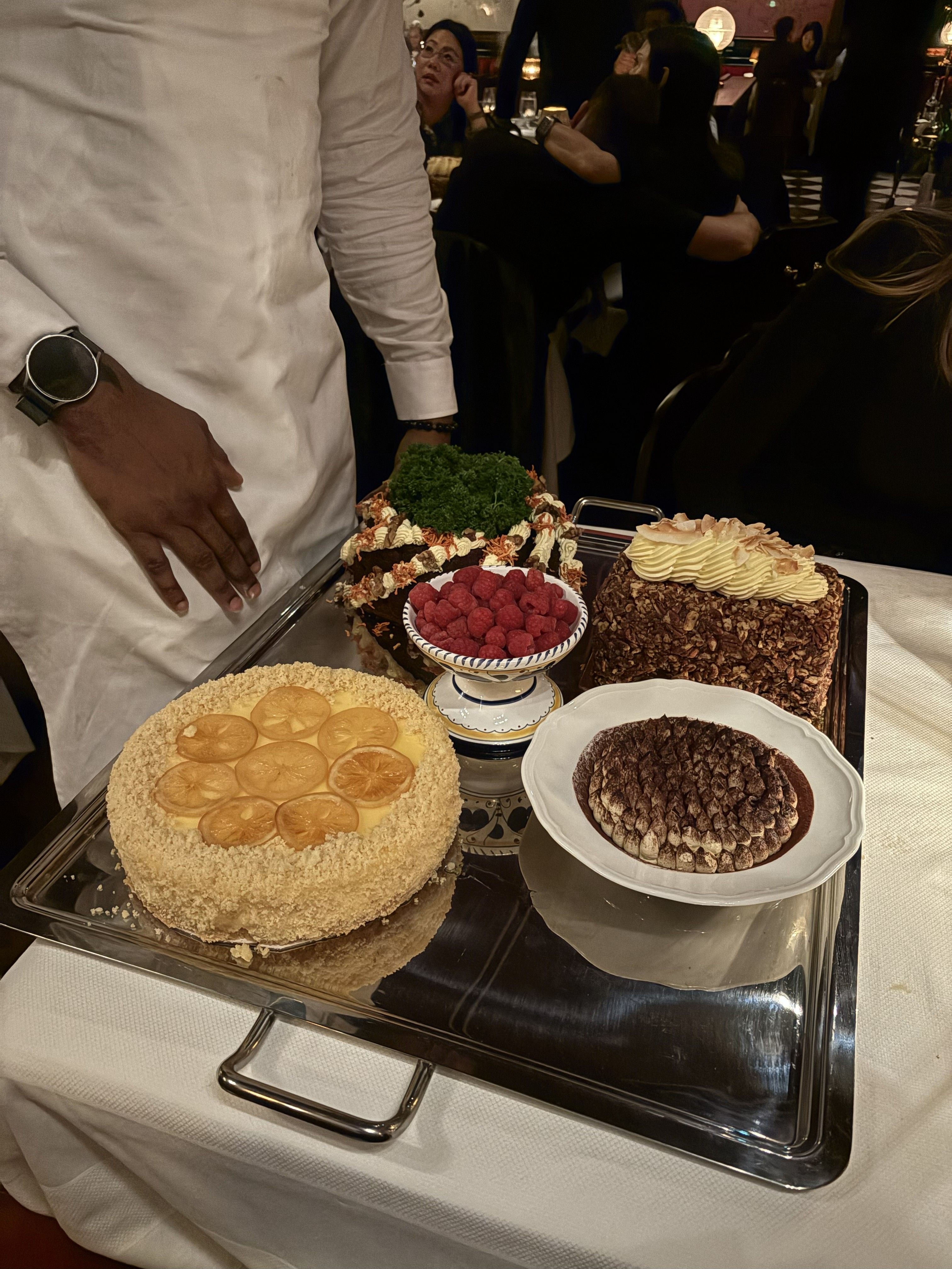 Three image, the first depicting a hand as it holds a double-A3 restaurant menu with a handsketched restaurant exterior on its cover, the other depicting a cake trolley filled with desserts, and another capturing a hand holding a white dish topped with silver sauce boats.