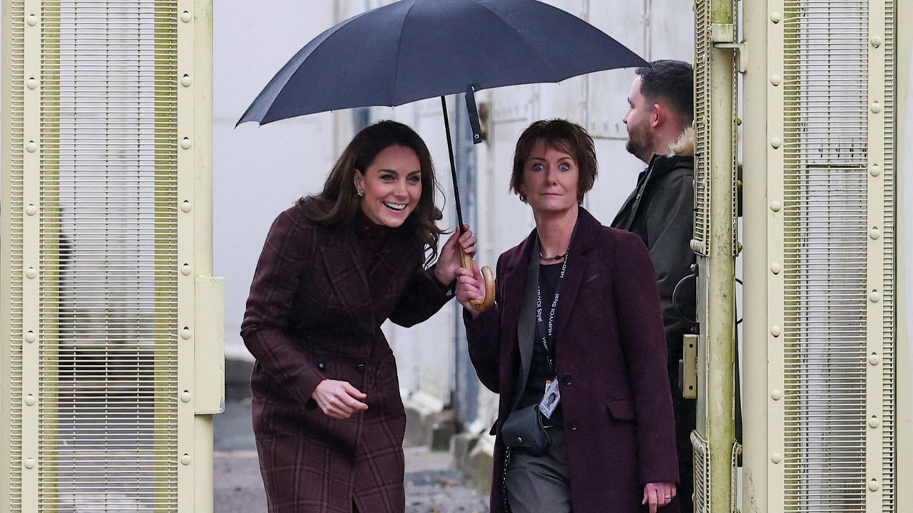 Kate Middleton walking through prison gates ducking under an umbrella that is being held by a woman in a blazer making a surprised expression