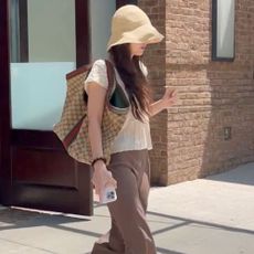 Dakota Johnson steps outside wearing dark brown slingback flats with brown wide-leg trousers and white lace t-shirt. She completed her look with a Gucci bag and straw bucket hat.