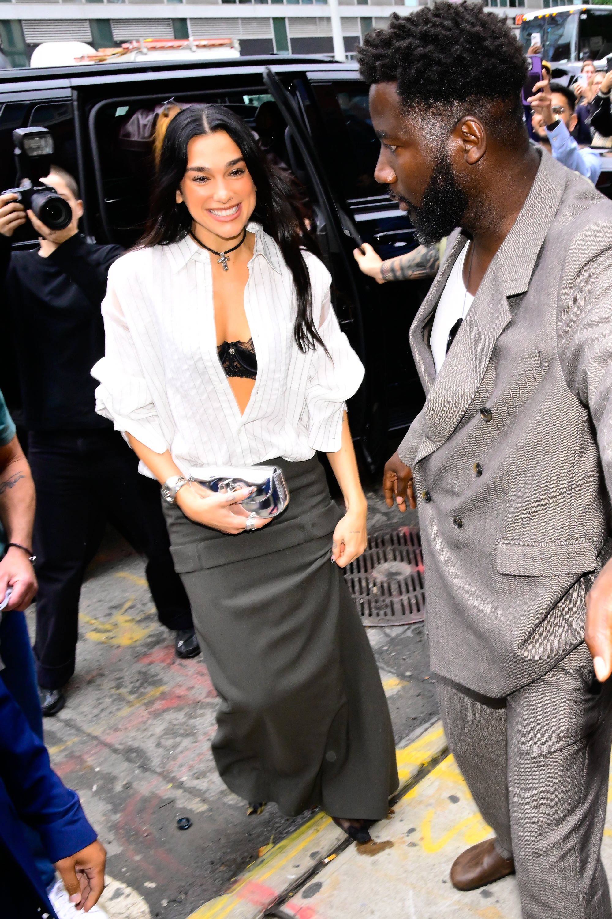 a woman with long dark hair wearing a white, striped button down shirt and a gray maxi skirt being photographed on the street by paparazzi