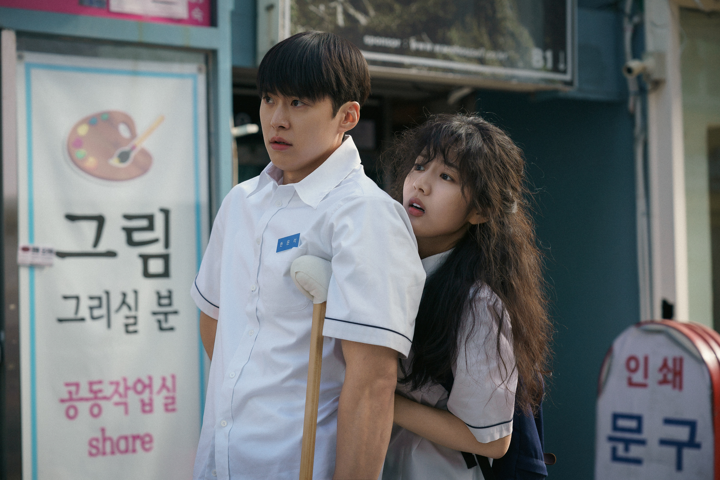 A high-school boy(Gong Myoung as Han Yun-seok) stands with crutches as a high-school girl (Shin Eun-soo as Park Se-ri) hides behind him, in the Netflix Korean movie 'Love Untangled.'