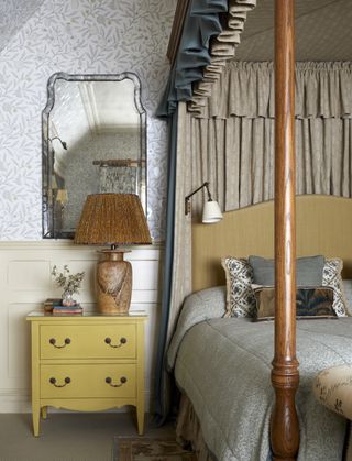 A bedroom with botanical wallpaper, a yellow side table, and a wooden four-poster bed frame