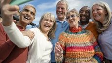 A group of senior friends/couples is looking at a smartphone while taking a selfie, laughing together.