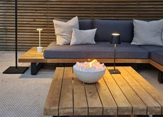 A backyard with a firepit bowl on a wooden slatted table with a grey sofa in the background