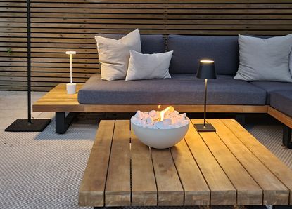 A backyard with a firepit bowl on a wooden slatted table with a grey sofa in the background