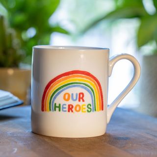 a white mug with a rainbow on it and the words 'our heroes' written on it, sitting on a wooden kitchen surface