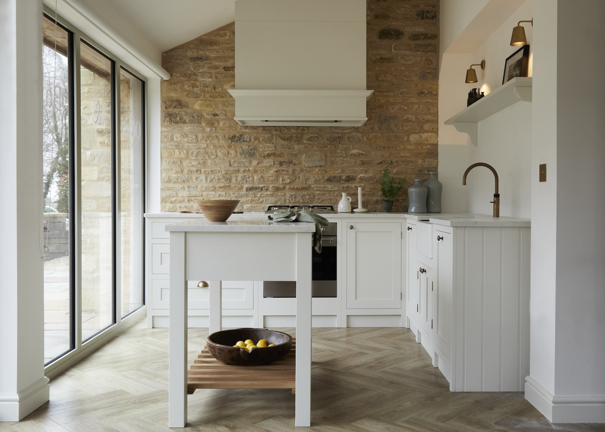 A white kitchen with a brick wall. In the middle of the small kitchen is a small island with open shelving at the bottom. There is a wooden bowl on top of the small island.