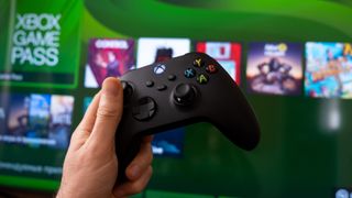 Hand holding Xbox Series X controller in front of a screen with Xbox Game Pass logo in background