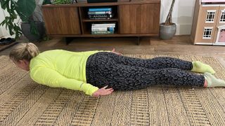 Kat Storr performs the prone cobra pose on a rug in a living room. She is wearing a long-sleeved top and leggings. She is face down on the floor, with her head and neck elevated. Her arms are by her sides and hover above thr ground. Behind her we see a leafy plant, TV cabinet and a doll's house.