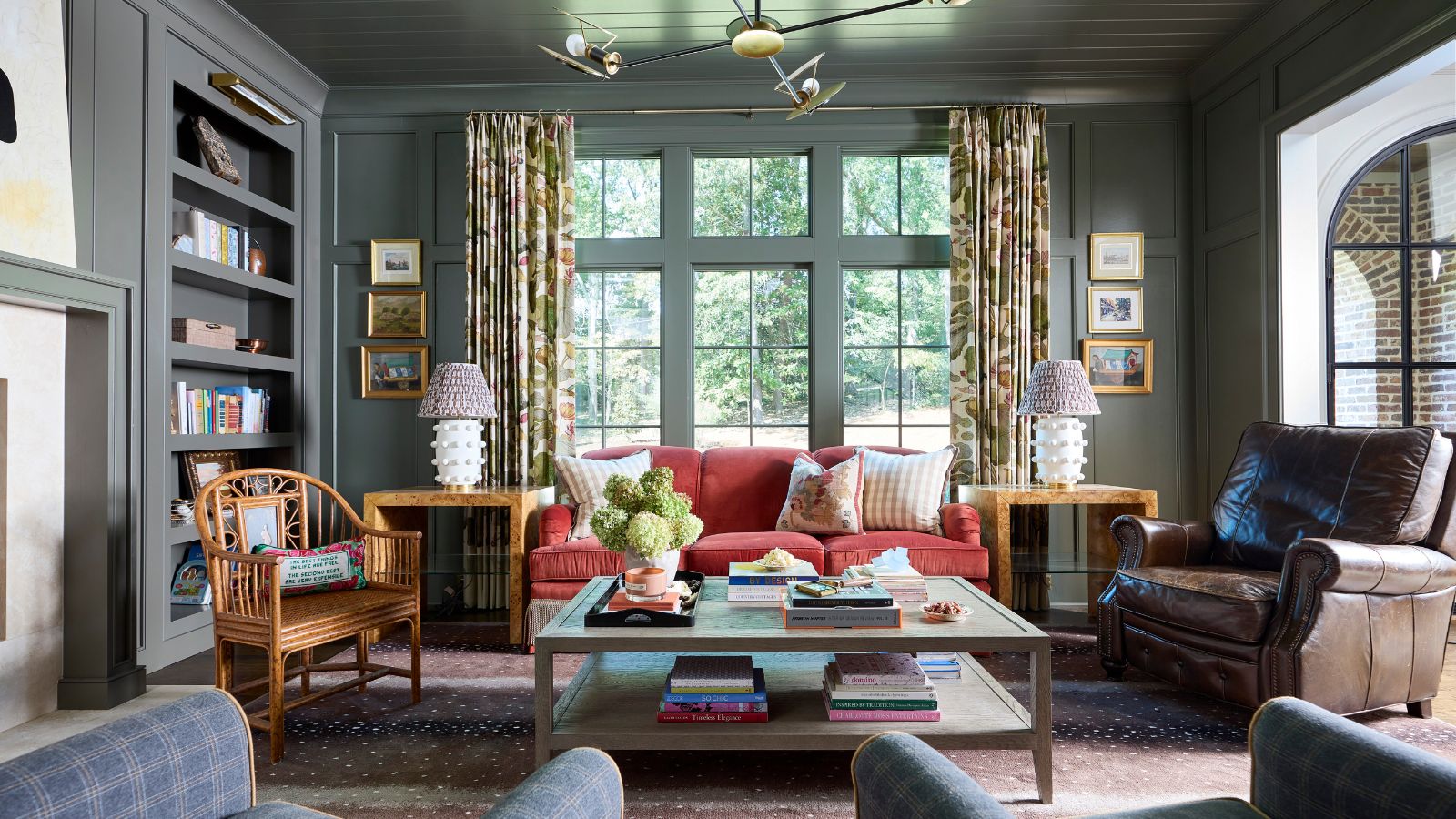 A living room with muted green color-drenched walls and ceiling, built-ins, a raspberry red sofa in front of the windows, and traditional decor. 