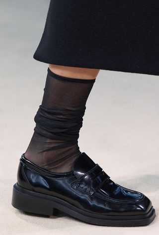 preppy loafer shoe trend is shown on cos's fall winter 2024 runway in a close photo of a black loafer paired with sheer stockings and a black skirt