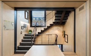 An interior view of part of a building featuring birch faced plywood panelling, with picture frames on the walls, black staircaseand a large window by the stairs with black boarders.