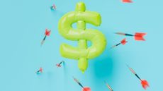 An inflated yellow dollar sign with darts being thrown at it. 