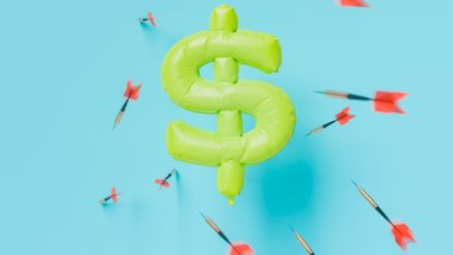 An inflated yellow dollar sign with darts being thrown at it. 