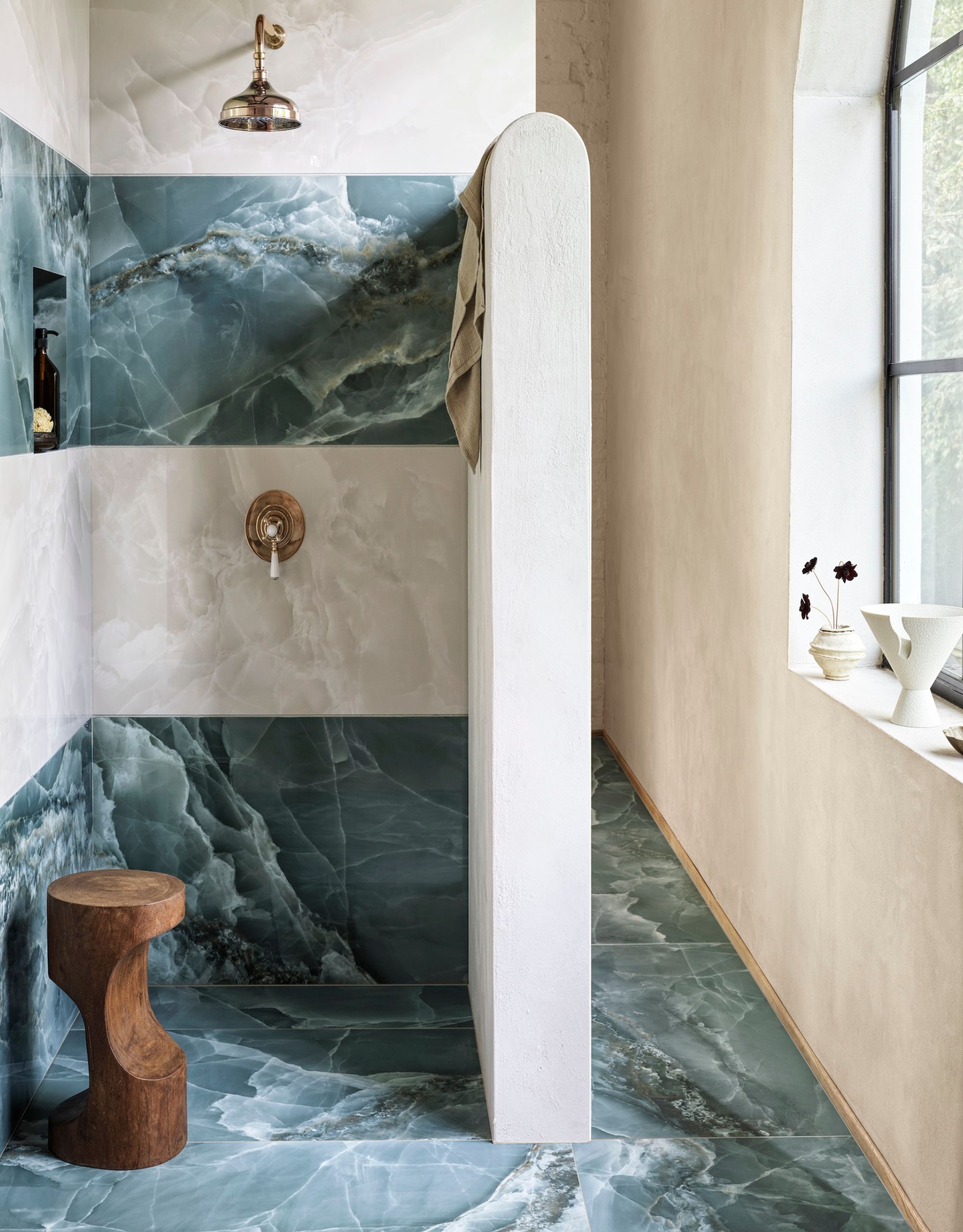 A walk-in shower with bands of alternating onyx tiles in blue and off-white for a striking look with brass details and a sculptural wooden stool