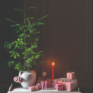 A sideboard with a potted miniature Christmas tree next to a pile of presents and a lit candle