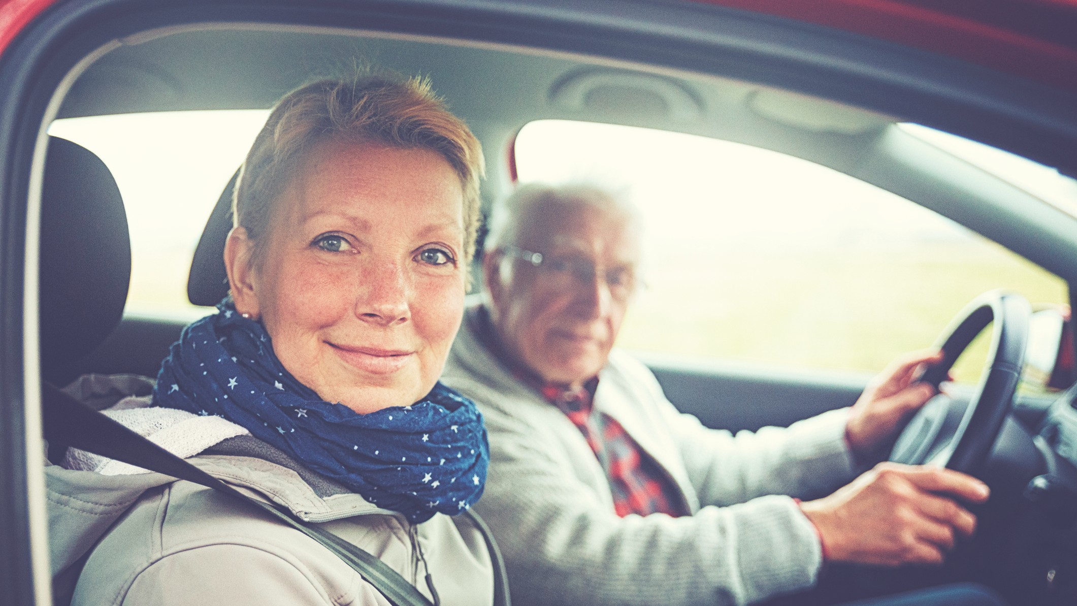 An older man drives his adult daughter. Both are looking at the camera and smiling.
