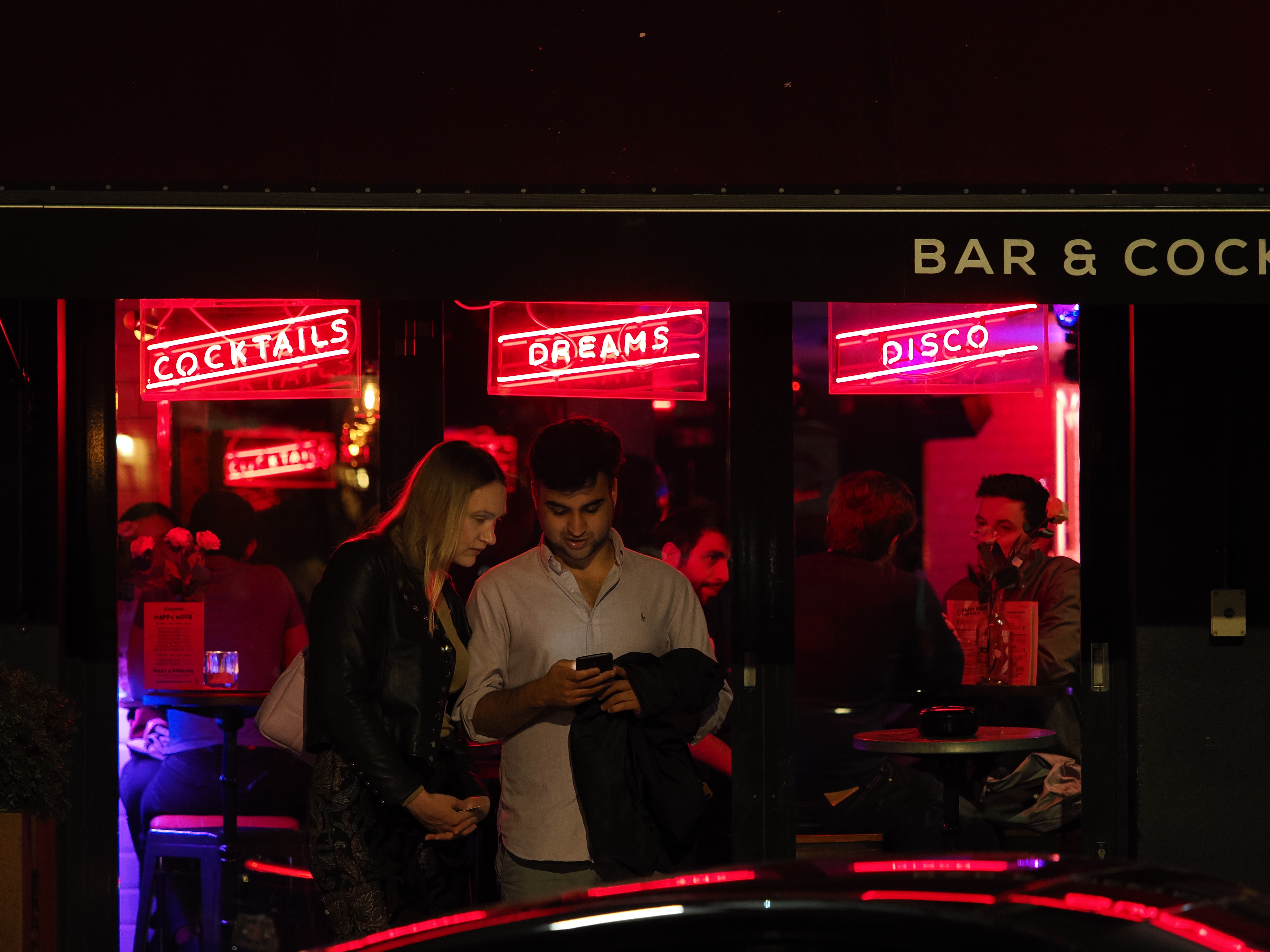 Two people standing outside a bar at night behind a taxi