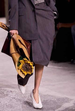 Woven shoe trend shown in Bottega Veneta's spring 2025 runway show with a model wearing a gray skirt suit set, a red handbag, white woven pumps, and a crochet sunflower