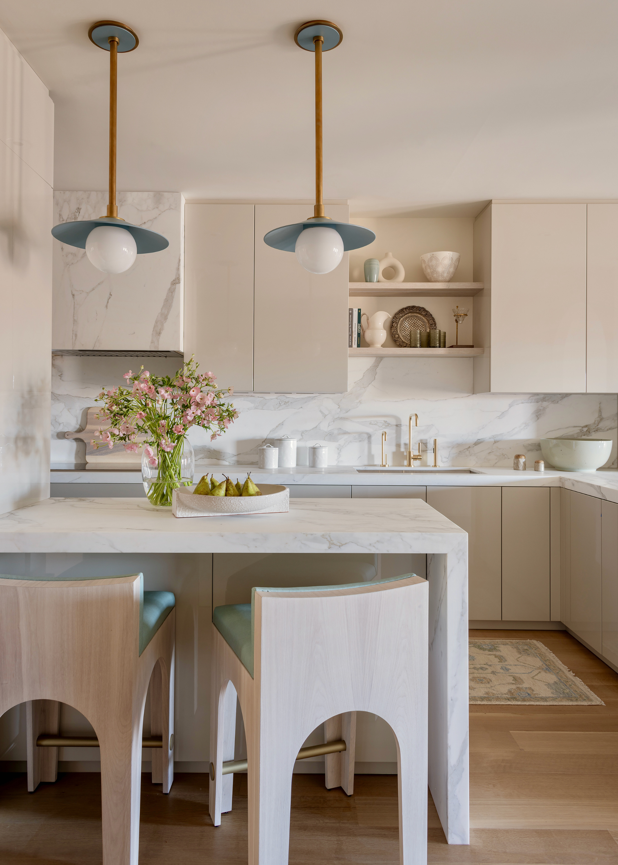 A modern neutral kitchen with marble backsplash, extractor hood and peninsula with cream cabinetry, pendant lighting and two bar stools with teal seat cushions