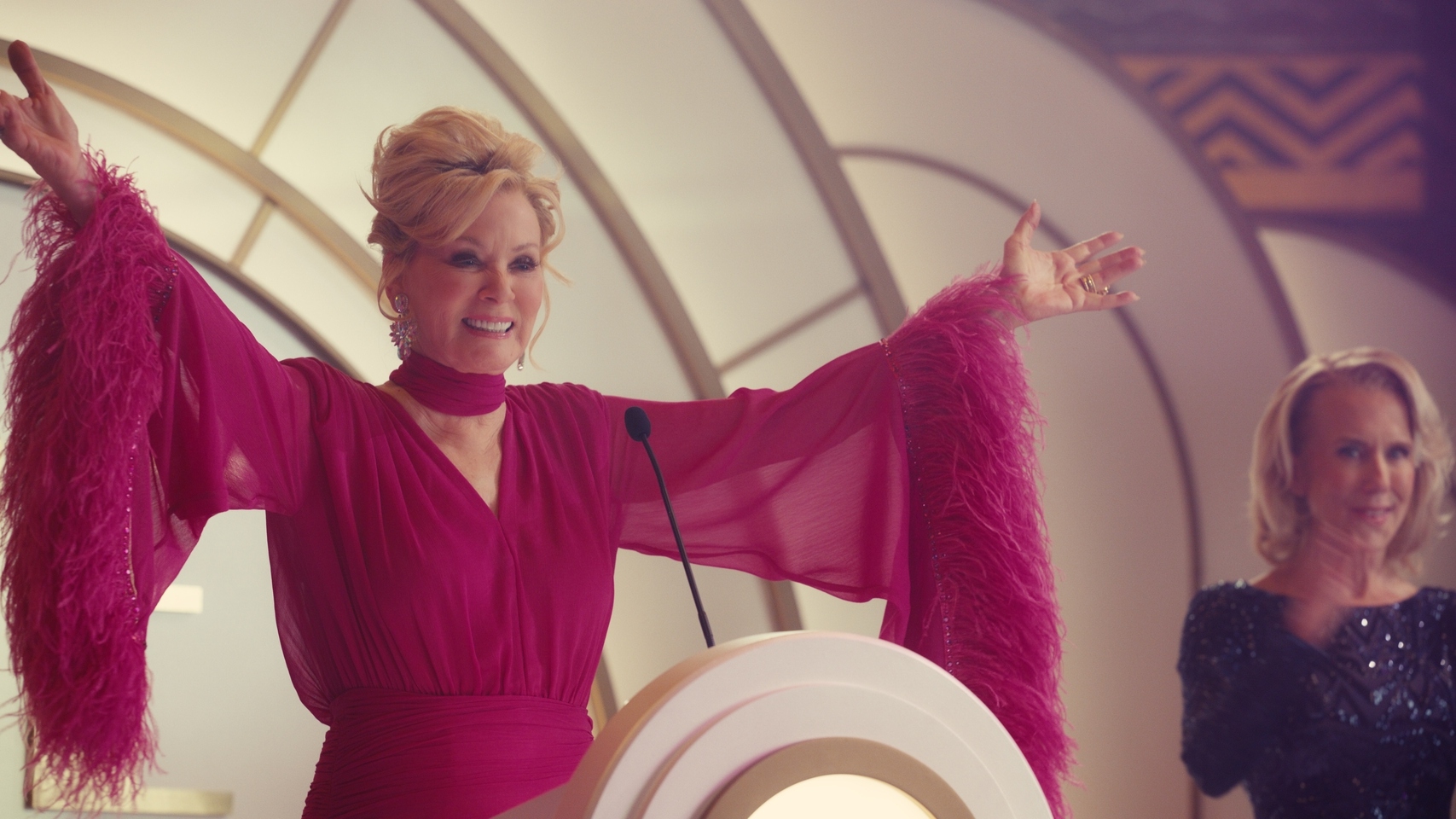 Deborah Vance as Jean with her arms out on stage wearing a pink dress with flare sleeves and feathers.