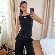 style influencer Ilana Torbiner poses for a mirror selfie in her hotel room at Château Voltaire in France wearing a black cord necklace with a silver bean pendant, a black boatneck tank top, thin black belt, and black pants