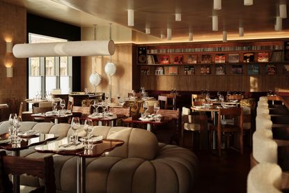 A retro-inspired restaurant dining room with oversized, modular sofas in beige, upholstered wooden bar stools in cream, wood-clad walls, sculptural lighting throughout, and a vinyl collection boldly displayed on its back wall.