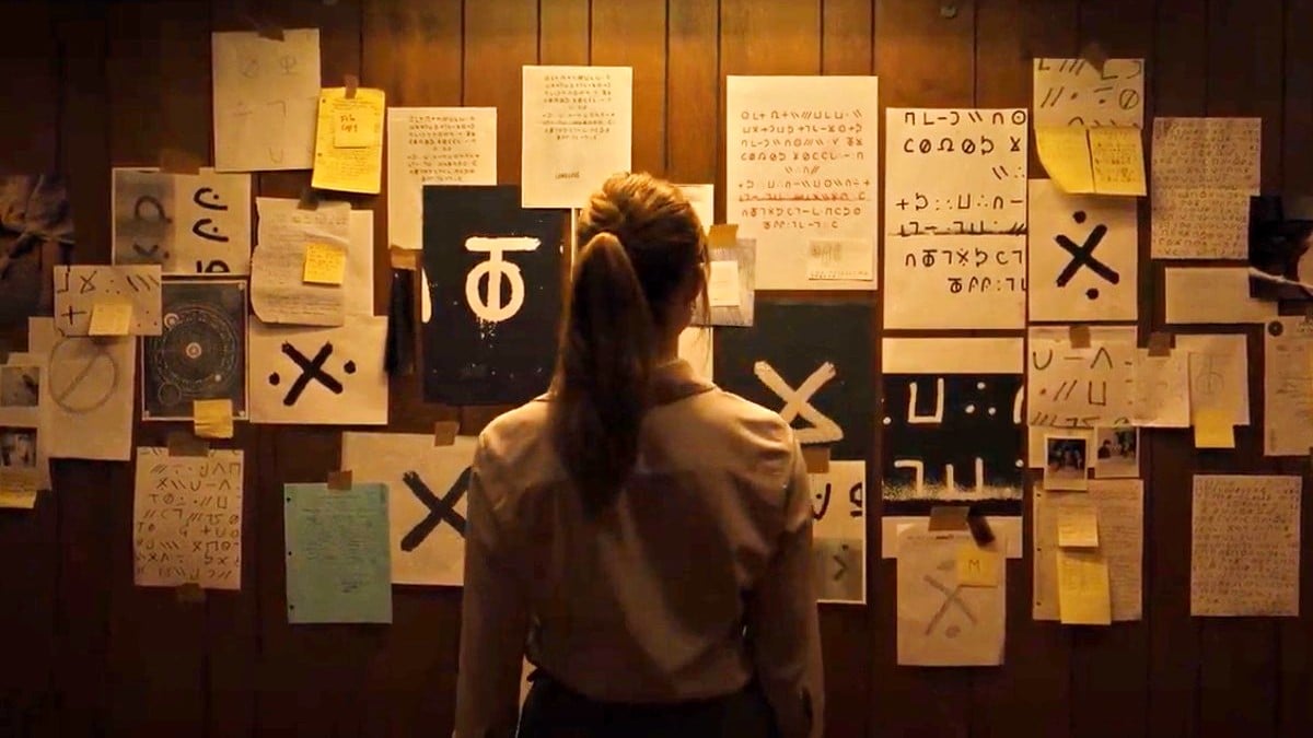 a woman stands in front of a wall covered with papers and cryptic drawings