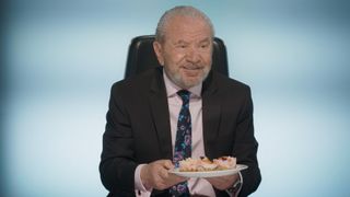 Lord Sugar holding a cheese cake and smiling.