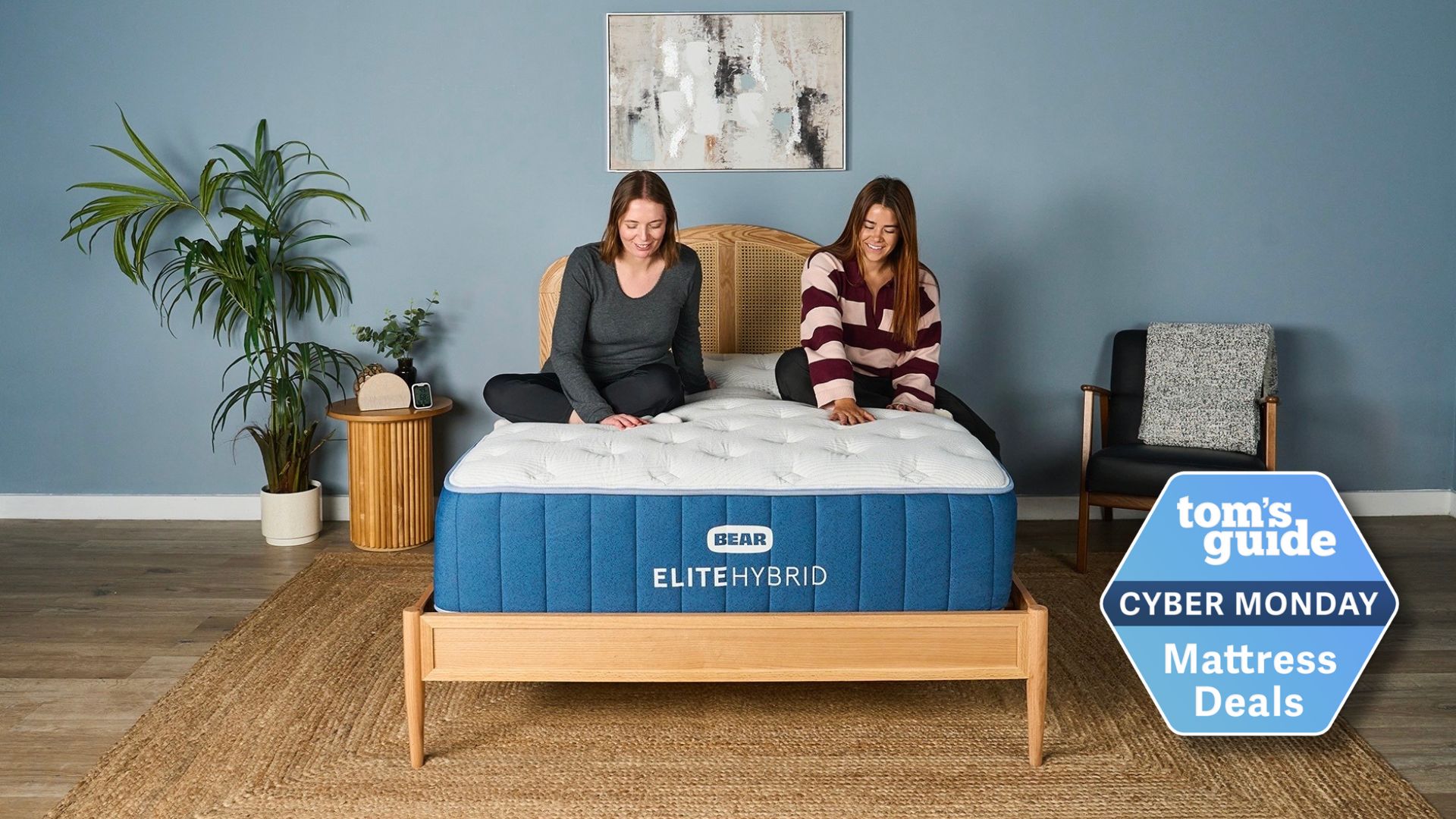 Two of our mattress testers sitting on the navy and white Bear Elite Hybrid mattress on wooden bedframe in sleep studio with blue wall, bedside chair, plants and Tom&#039;s Guide Cyber Monday mattress deals logo