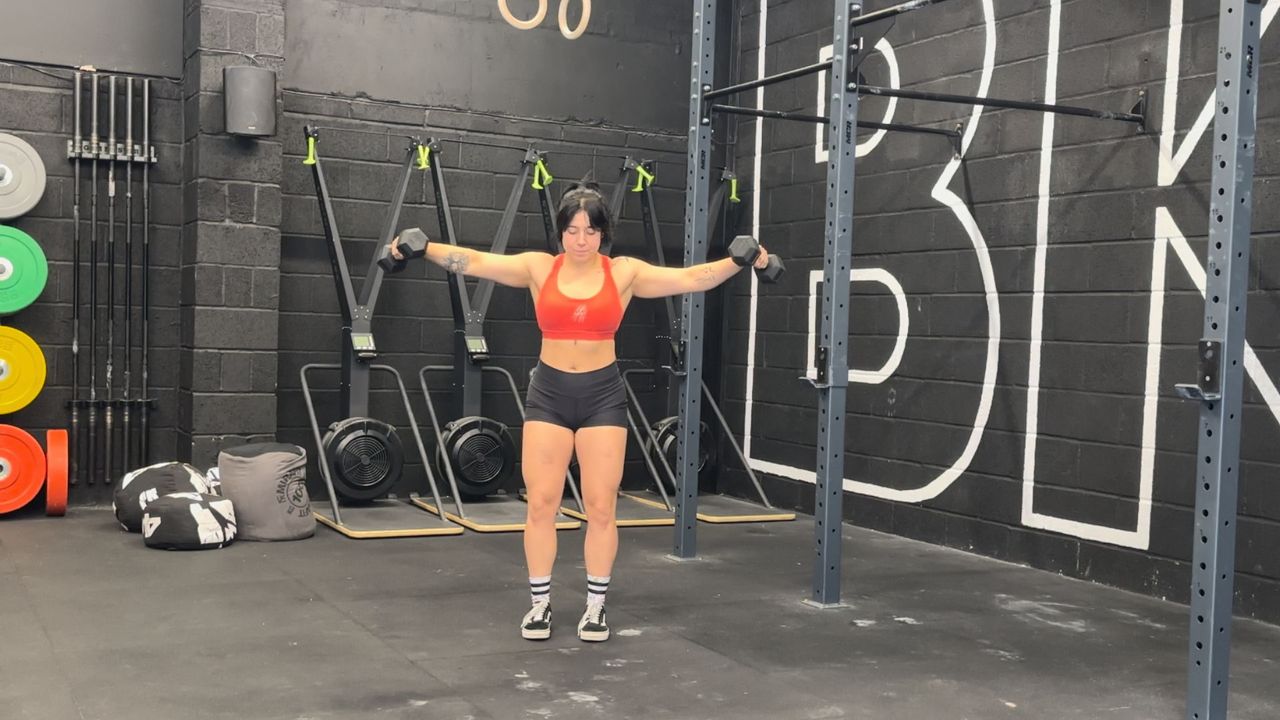 Raquel Sanjurjo performs a lateral to front raise. She is standing in a gym holding a pair of dumbbells out the sides, with her arms straight.