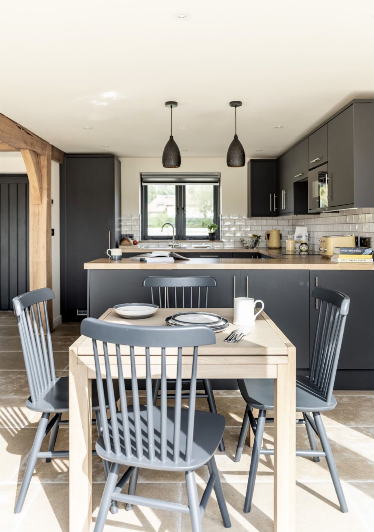A contemporary kitchen-diner with dark grey fittings, dark grey pendant lights, a white gloss subway tile backsplash and a wooden dinner table