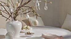 a vase of blooming branches decorated with Easter ornaments on a white tablecloth with napkins and bowls of food