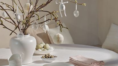a vase of blooming branches decorated with Easter ornaments on a white tablecloth with napkins and bowls of food