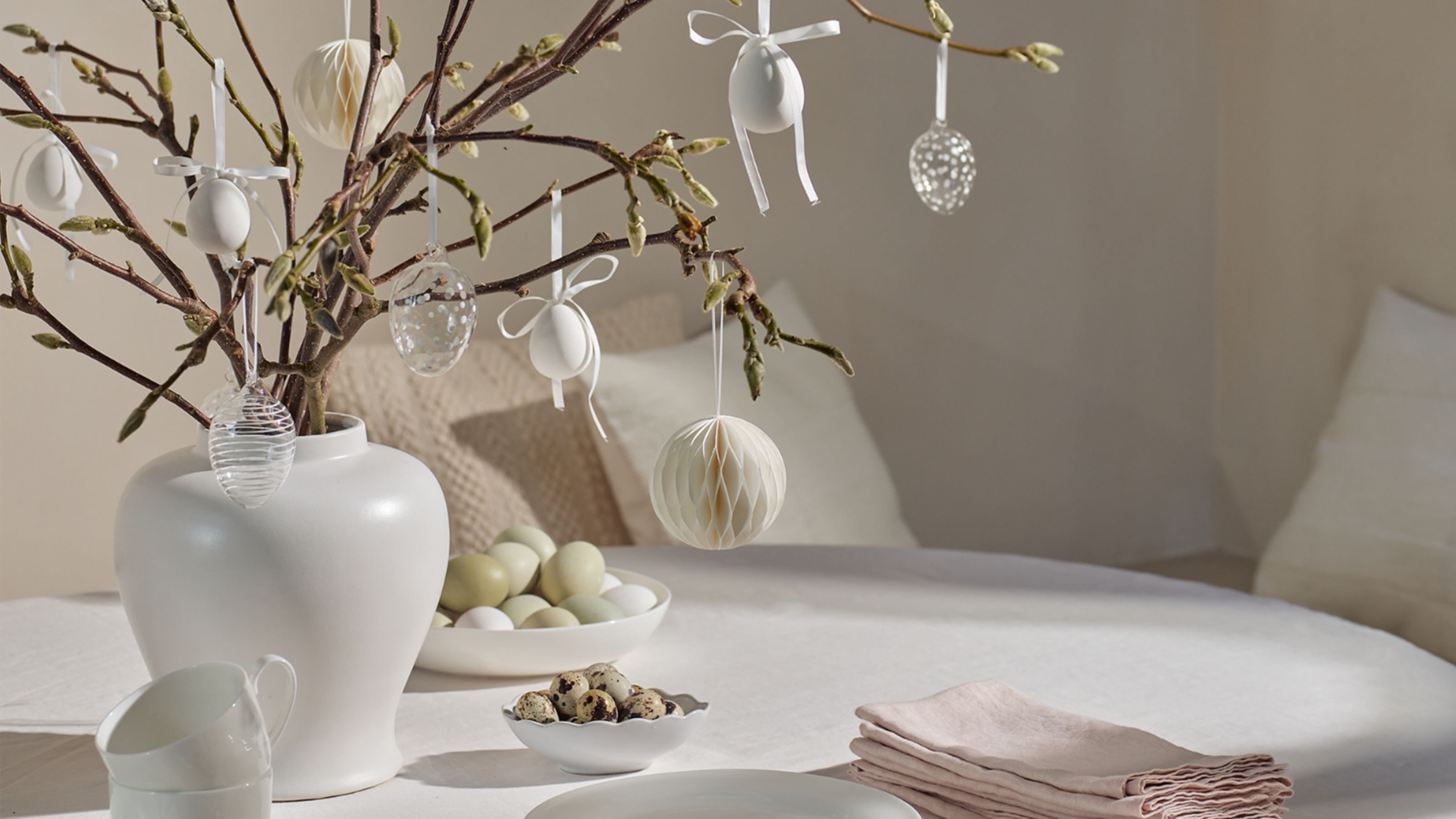 a vase of blooming branches decorated with Easter ornaments on a white tablecloth with napkins and bowls of food
