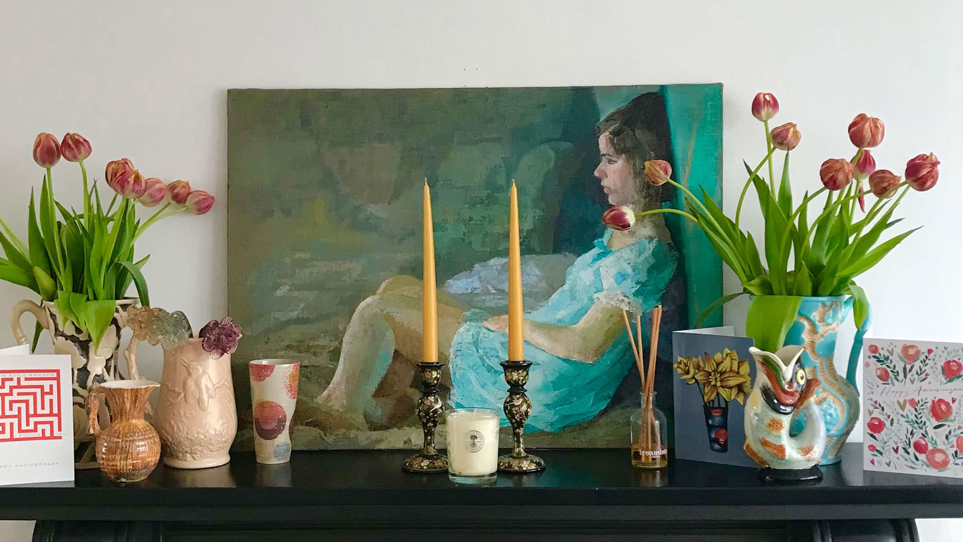 Fireplace mantlepiece with portrait of a girl and displaying candlesticks, cards and vases of tulips