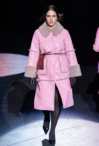 One of the best jacket coat brands, Baum und Pferdgarten, is shown in an image of a model wearing a pink shearling coat with black tights, black leather gloves, and black pumps