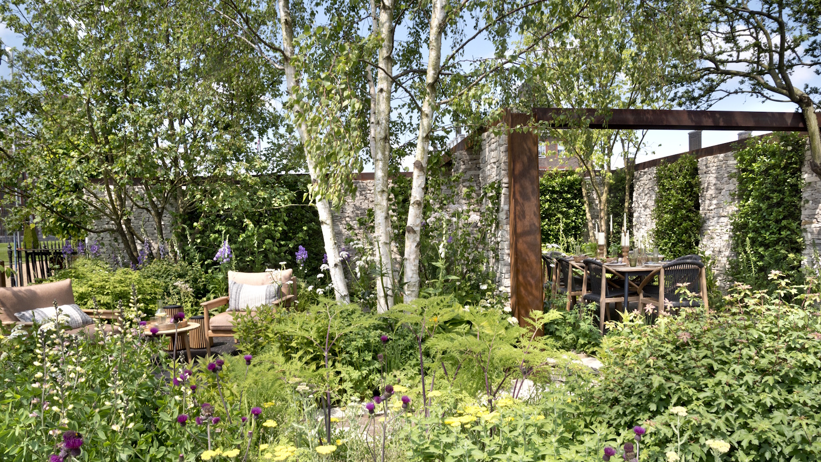 Lush garden with outdoor armchairs, a Corten steel pergola structure with dining table, under canopy of tall silver birch tree