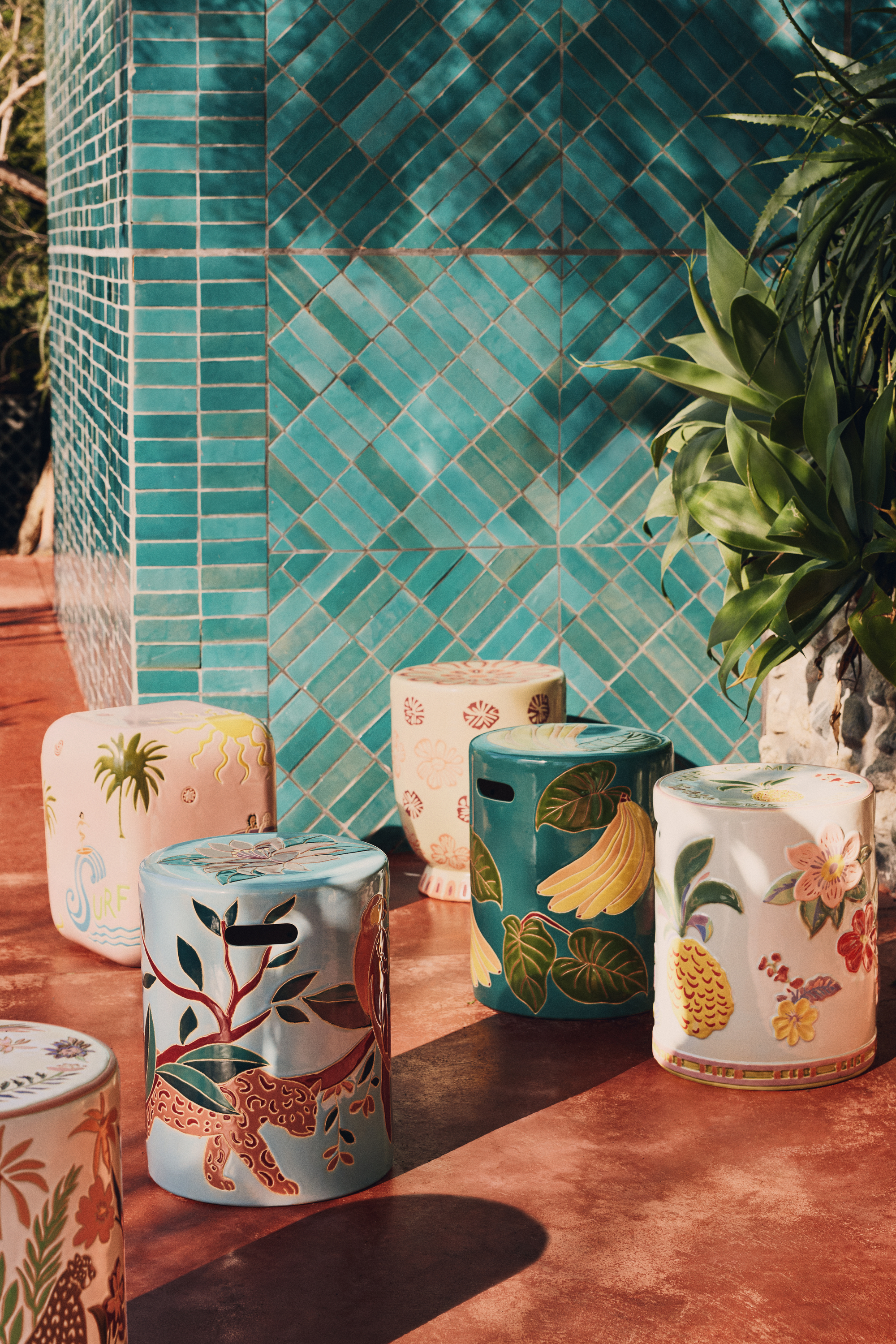 Image of six different colorful, ceramic, patterned side table on a terracotta deck with the wall of a house that is covered in teal tiles.