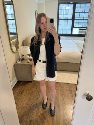 Nikki Chwatt wears Agolde Dame bermuda shorts in white with a black belt, black ballet flats, a white tank top, and a black button-down shirt.