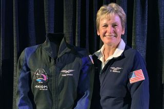 a smiling woman in a blue jacket stands next to a dark blue flight suit.
