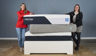 Two members of our sleep team stand either side of a stack of the best memory foam mattresses we've tested, including the Nectar and Siena, balanced on a bed frame